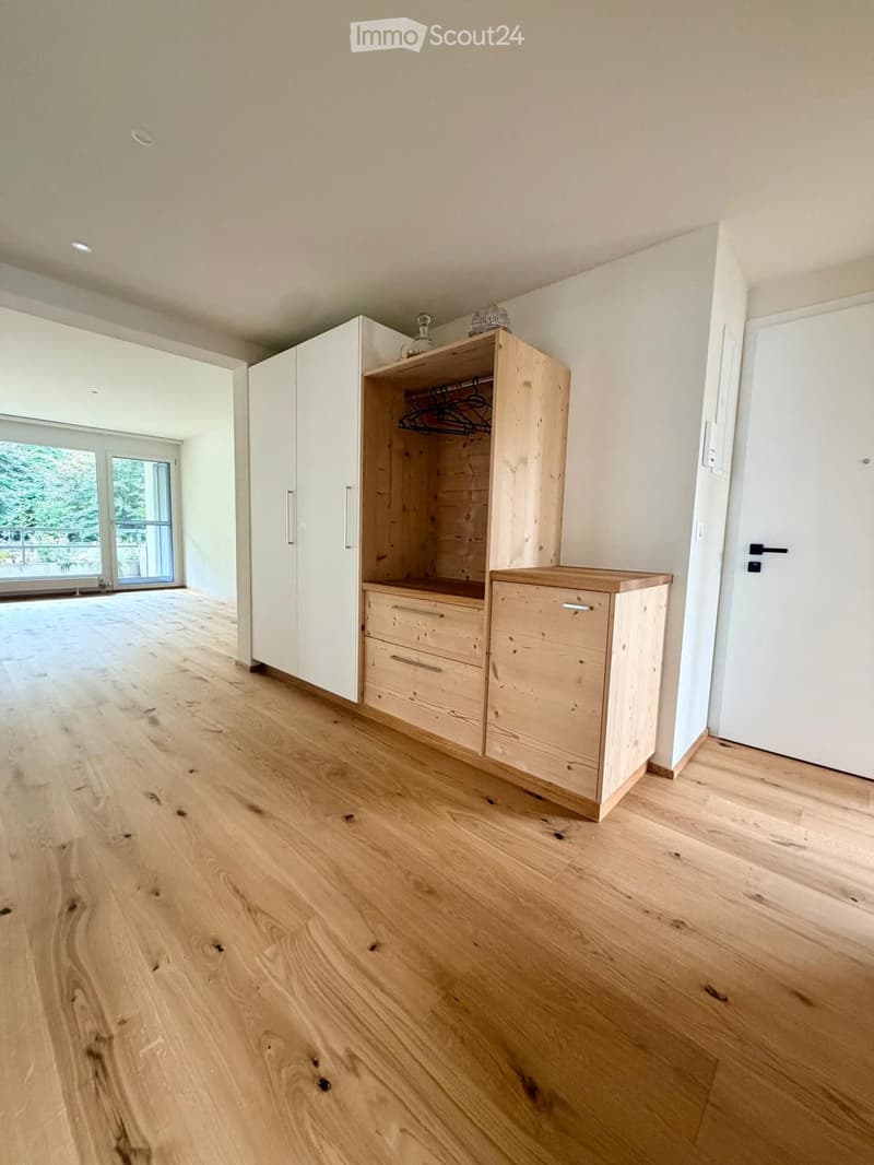 Empty room with light wooden flooring, white walls, built-in wooden closet with shelves and drawers, and large windows with a view of greenery.