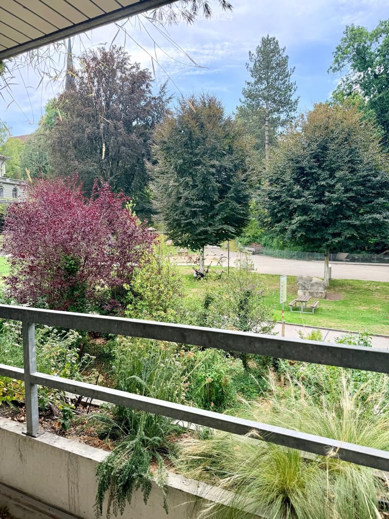 Balcony overlooking a park with trees, grass, a picnic table, and a stone structure.