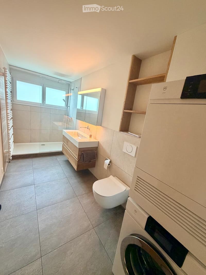 Modern bathroom with a sink, mirror, toilet, shower, and a washing machine. Features include wooden cabinets, tiled floors, and large windows.