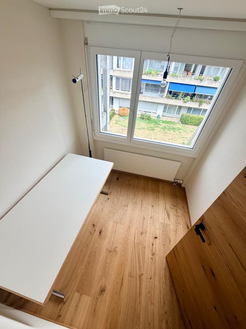 Empty room with wooden flooring, white walls, a large window with a view of a garden and neighboring building, a radiator, and a small table near the window.