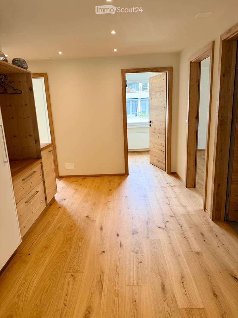 Empty hallway with light wooden flooring, white walls, and recessed lighting. Features a wooden wardrobe with drawers and a mirror, and an open doorway leading to another room.