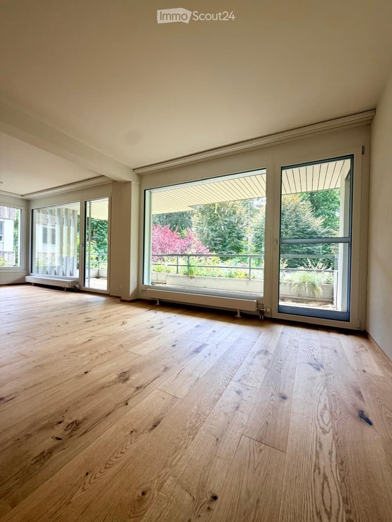 Empty room with light wooden flooring, large windows, and sliding glass doors leading to a balcony area with outdoor plants.