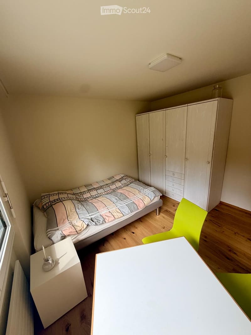 A small, furnished bedroom with a single bed, a green chair, a white table, and a large white wardrobe. The room features wooden flooring and a window on the left side.