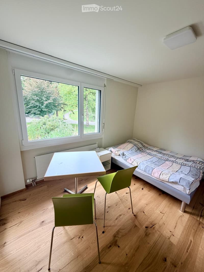 A small, furnished room with a bed, a table, and two chairs, featuring wooden flooring and large windows overlooking a green area.