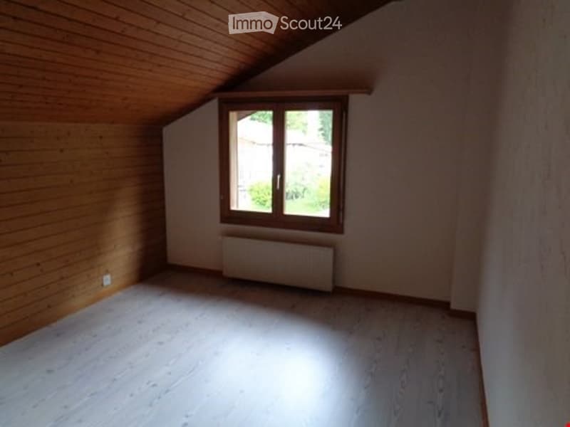 Empty attic room with wooden walls and floor, single window, radiator, and exposed wooden beams on the ceiling.