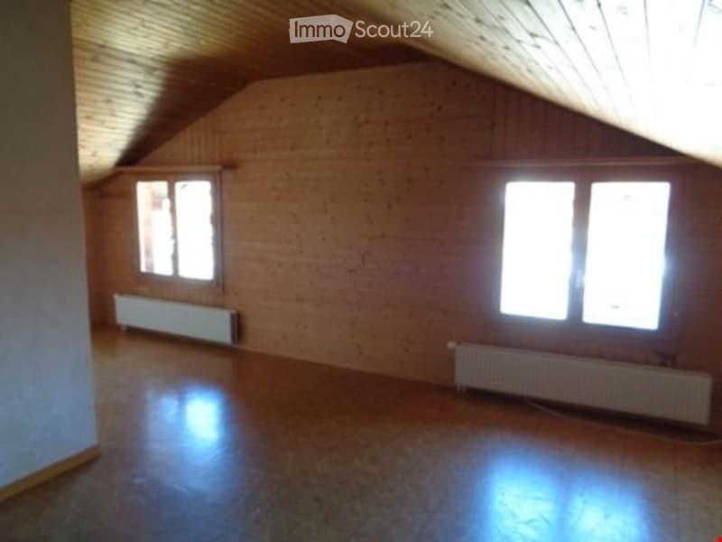 Empty attic room with wooden walls, slanted wooden ceiling, and two windows. The floor is polished wood, and there are radiators beneath the windows.