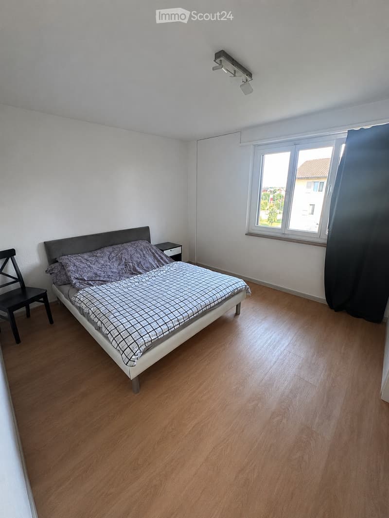 Bedroom with wooden flooring, a bed with a grey headboard and patterned bedding, a nightstand, and a chair near the window.