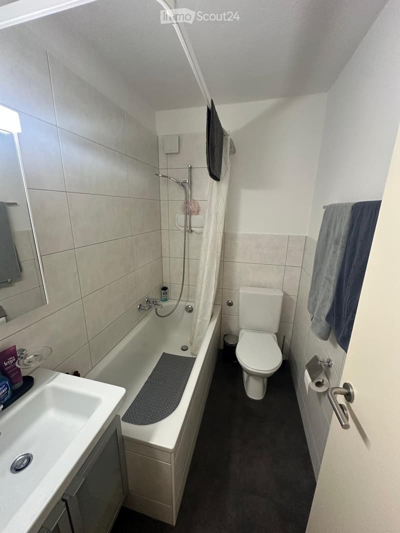 Modern bathroom with a white sink, bathtub, toilet, and mirror. Towels are hanging on the wall, and a small shelf holds cleaning supplies.