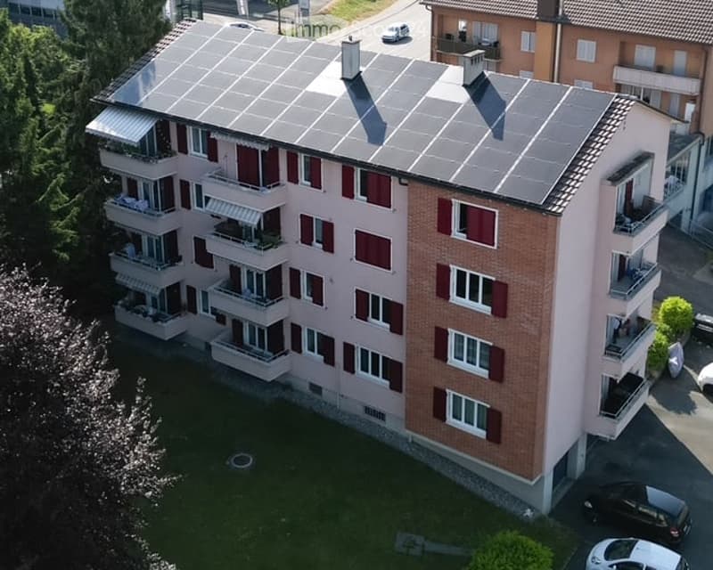 Aerial view of a multi-story residential building with solar panels on the roof, balconies, and surrounding greenery.