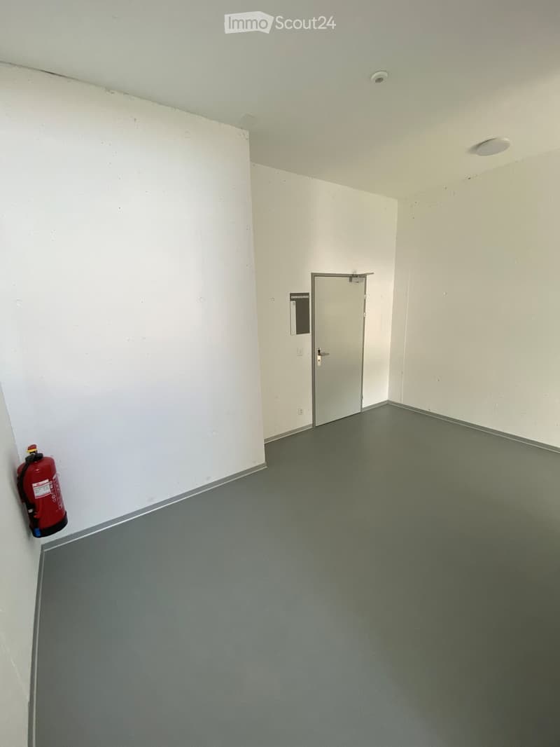 Empty room with white walls, grey floor, and a single door leading to another space. A red fire extinguisher is mounted on the wall.