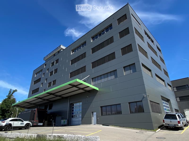Modern multi-story building with grey facade, large windows, green awning over entrance, and parking area with vehicles.