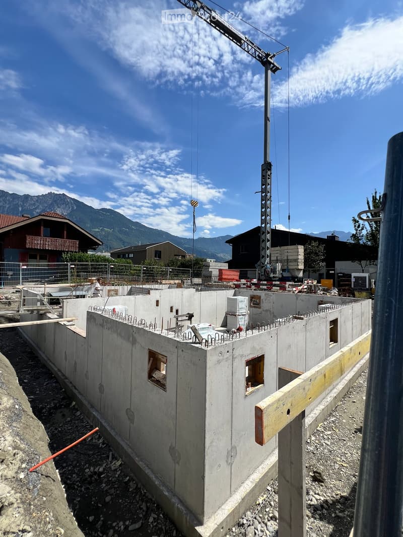 A partially constructed building with concrete walls, exposed rebar, and a crane in the background, set against a mountainous backdrop.