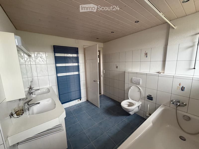 Bathroom with two sinks, a toilet, a bathtub, and a shower area. The room features white tiled walls, blue tiled flooring, and a window with blue blinds.