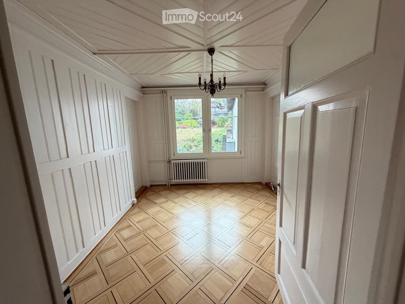 Empty hallway with wooden herringbone flooring, white paneled walls, and a chandelier hanging from the ceiling. Natural light streams in from a window at the end of the corridor.