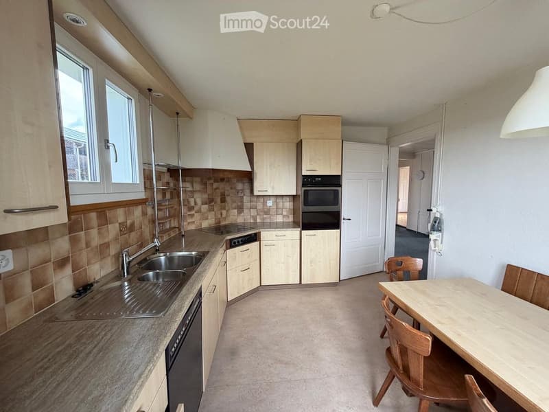 Modern kitchen with light wood cabinets, stainless steel sink, oven, dishwasher, and a small dining area with a table and chairs.