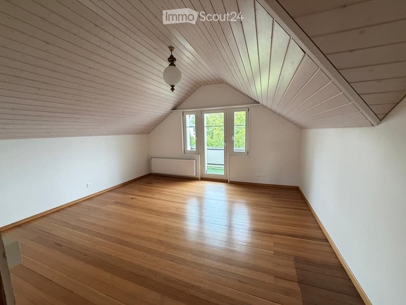 Empty attic room with wooden floors, white walls, and a slanted wooden ceiling. Features a single window and a door leading outside.