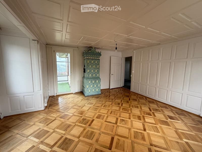An empty room with wooden parquet flooring, white paneled walls, and a decorative green cabinet in the center. The room has several doors, including one leading to a balcony with a green outdoor area visible.