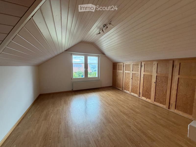 Empty attic room with wooden flooring, white walls, built-in wooden closet, and a large window offering a view of the outside.