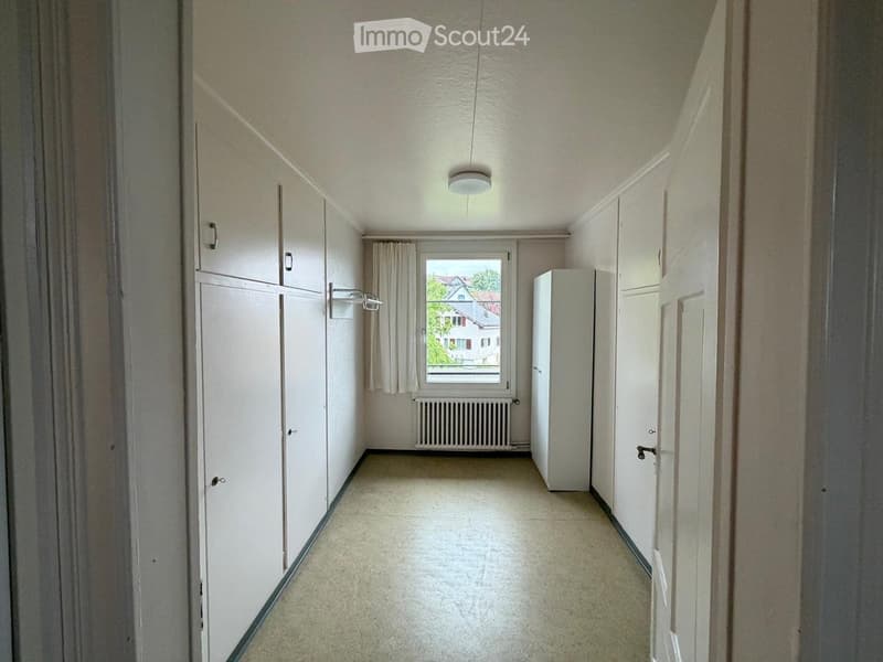 Empty hallway with white walls, beige carpet, built-in wardrobes, a window with a view of a house, and a radiator.