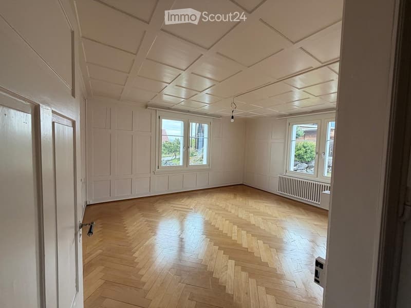 Empty room with wooden herringbone flooring, white paneled walls, large windows, and a radiator. The ceiling features a decorative panel design with recessed lighting.