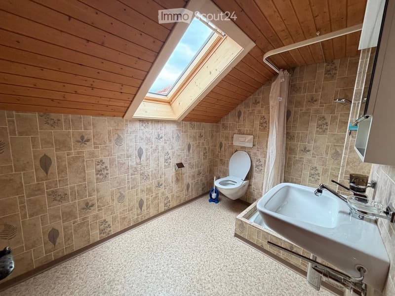 Cozy attic bathroom with wooden ceiling, skylight window, tiled walls, toilet, sink, and mirror.