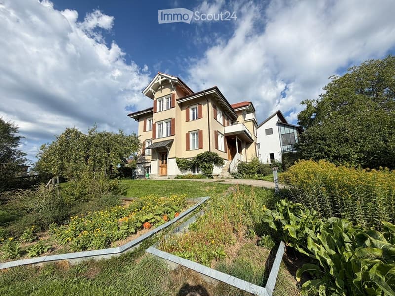 A two-story house with a beige facade, red shutters, and a well-maintained garden in front.