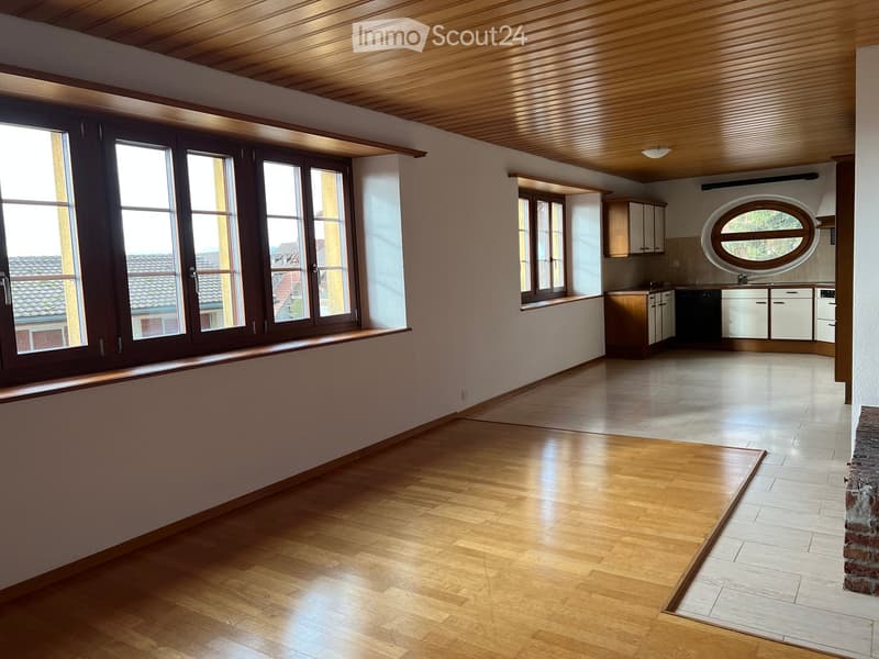 Bright, open living space with wooden flooring, large windows, and a small kitchen area featuring white cabinets and a dishwasher.