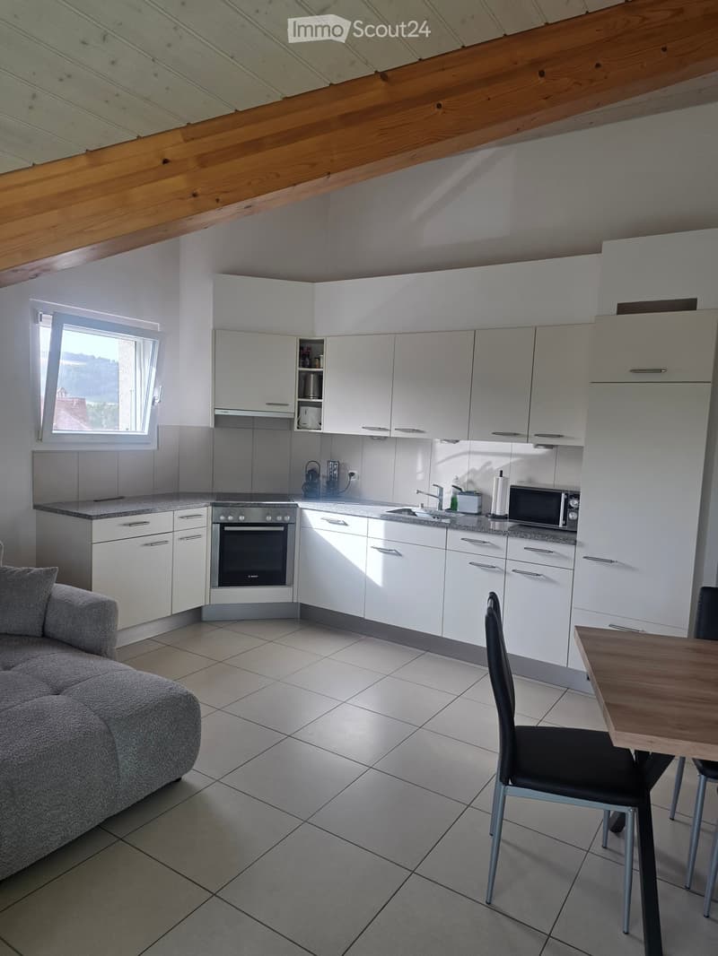 Modern kitchen with white cabinets, stainless steel appliances, and a grey sofa in the foreground. A dining table and chairs are also visible.