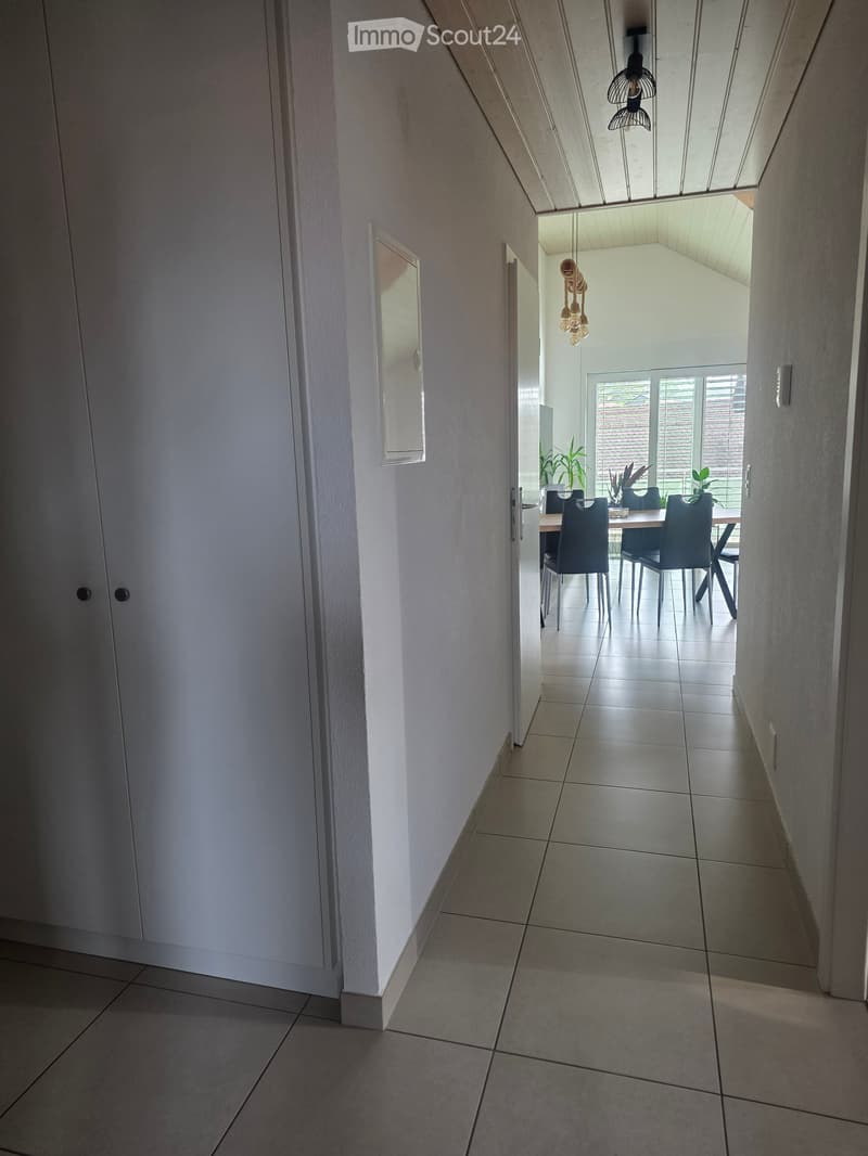A modern hallway with tiled flooring, white walls, and a closed door on the left. The hallway leads to a dining area with a table, chairs, and plants visible in the background.