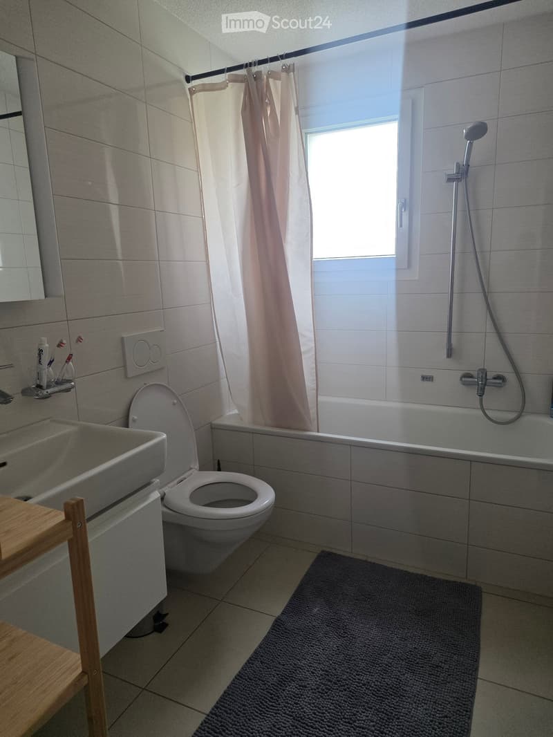 Modern bathroom with a toilet, sink, bathtub, and shower. Features a grey tiled wall, a window with a shower curtain, and a small wooden shelf holding toothbrushes and toothpaste.