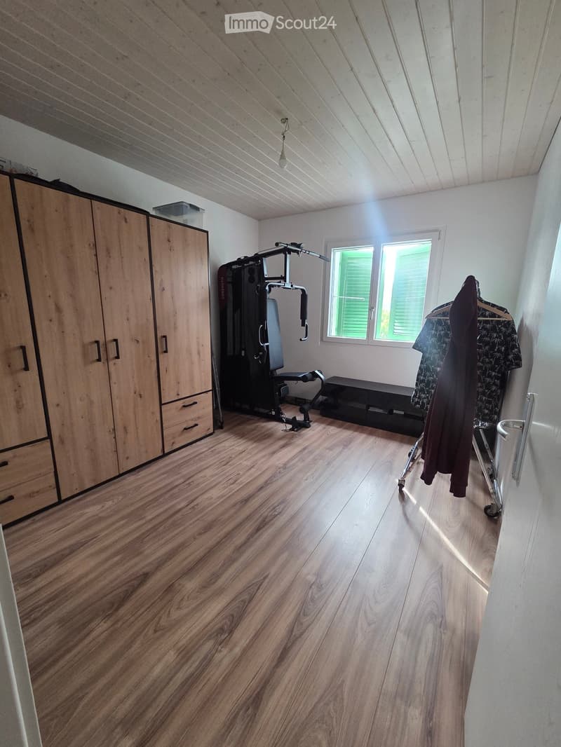A well-lit room with wooden flooring, a large wardrobe, an exercise bike, a bench, and a clothing rack with clothes hanging on it.