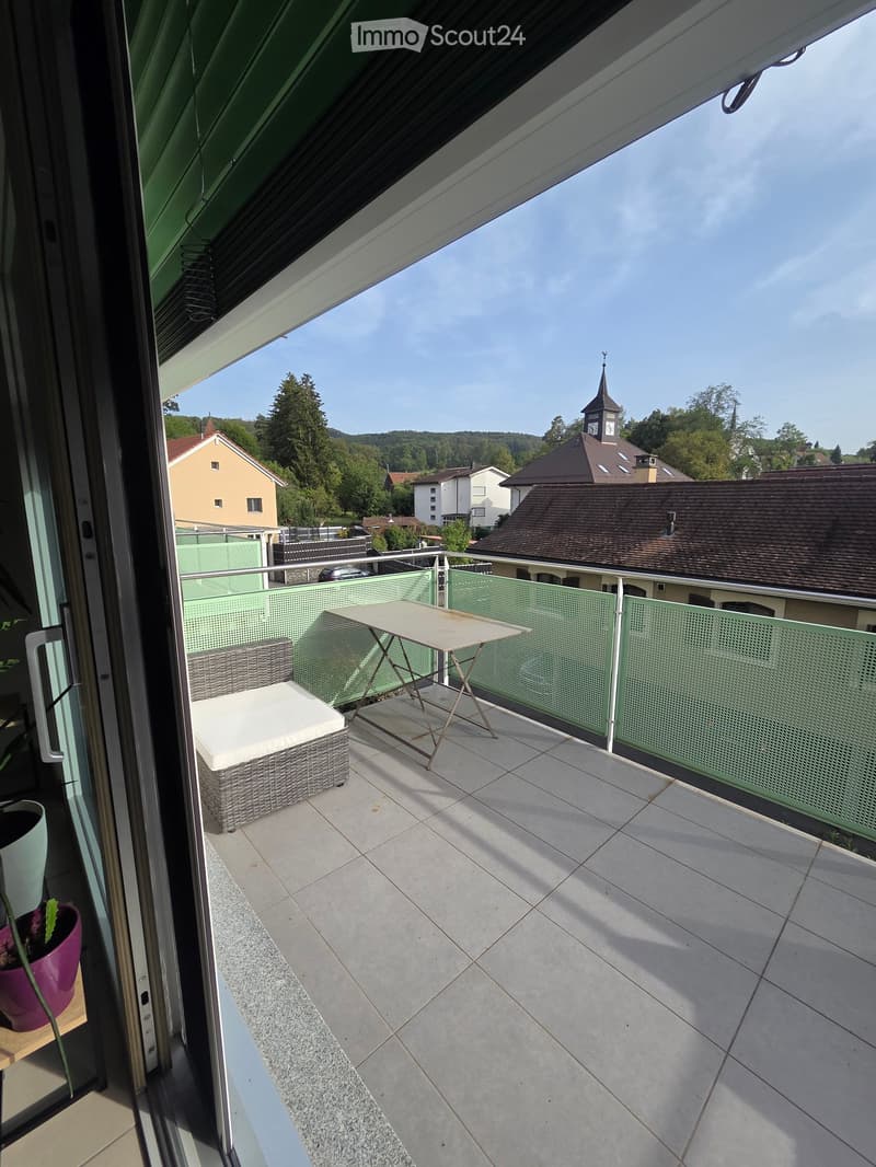 Balcony with tiled floor, green railing, small table, and wicker chair, overlooking a town with a church tower.