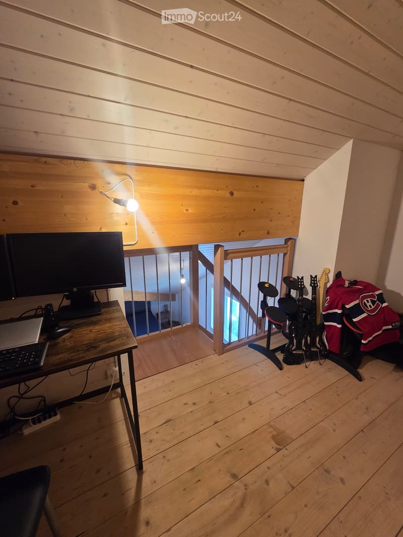 A wooden-floored room with a desk setup including a laptop, keyboard, and mouse, along with a drum set and guitars in the corner.