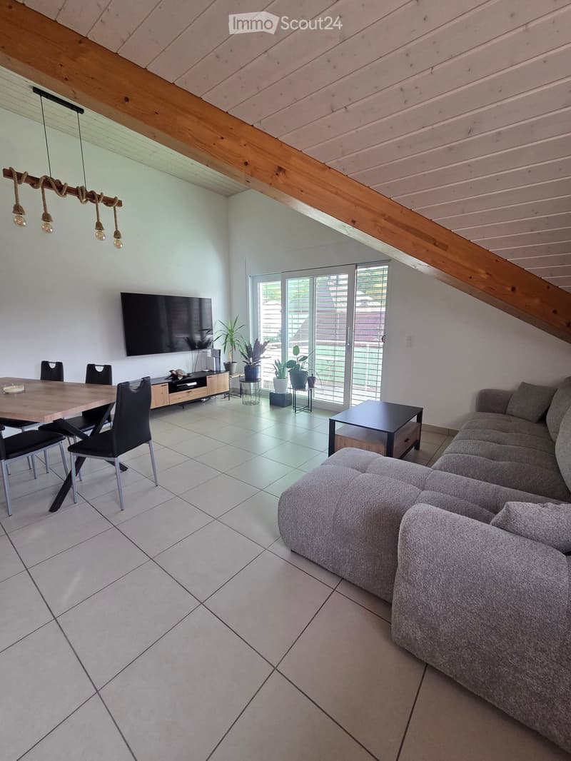 Modern living and dining area with a grey sectional sofa, coffee table, dining table with chairs, and a television mounted on the wall. The space features exposed wooden beams and a large sliding glass door leading to an outdoor area.