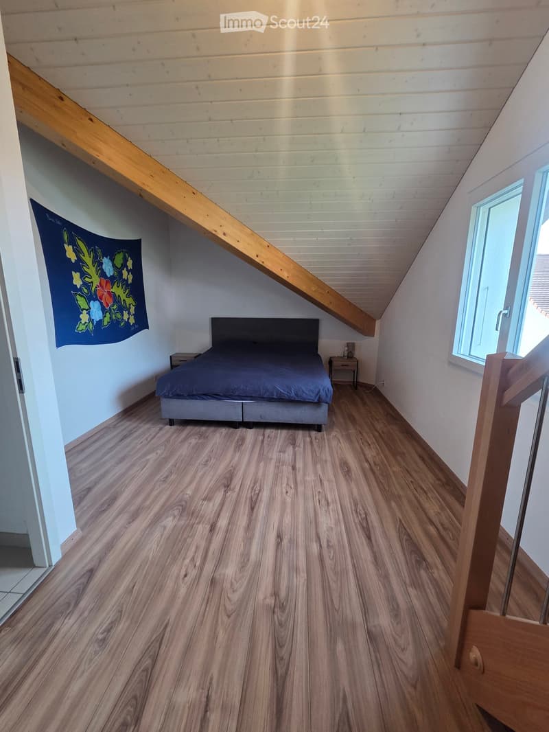 Cozy attic bedroom with wooden floors, slanted ceiling, large window, bed with dark headboard, nightstand, and decorative banner on wall.