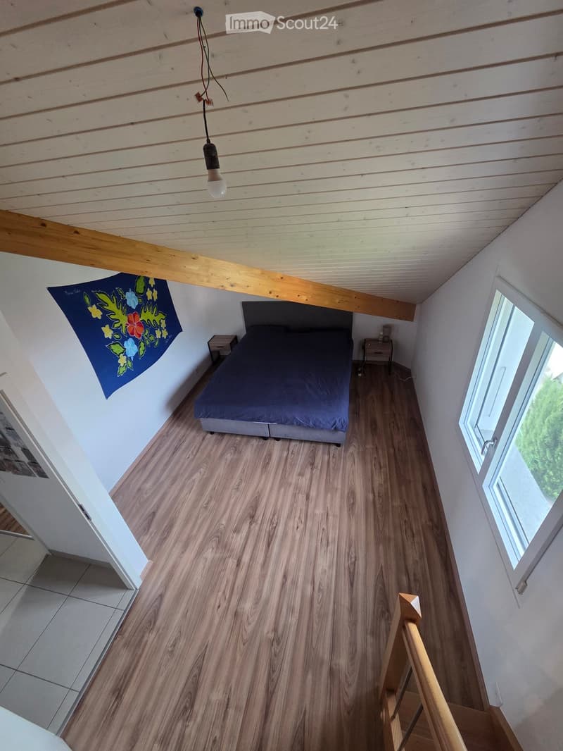 Cozy bedroom with wooden flooring, slanted wooden ceiling, bed with dark blue bedding, nightstands, and a hanging light fixture.