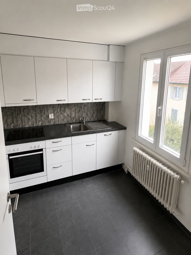 Modern kitchen with white cabinets, black countertops, stainless steel sink, and built-in oven. Dark tiled floor and radiator visible.