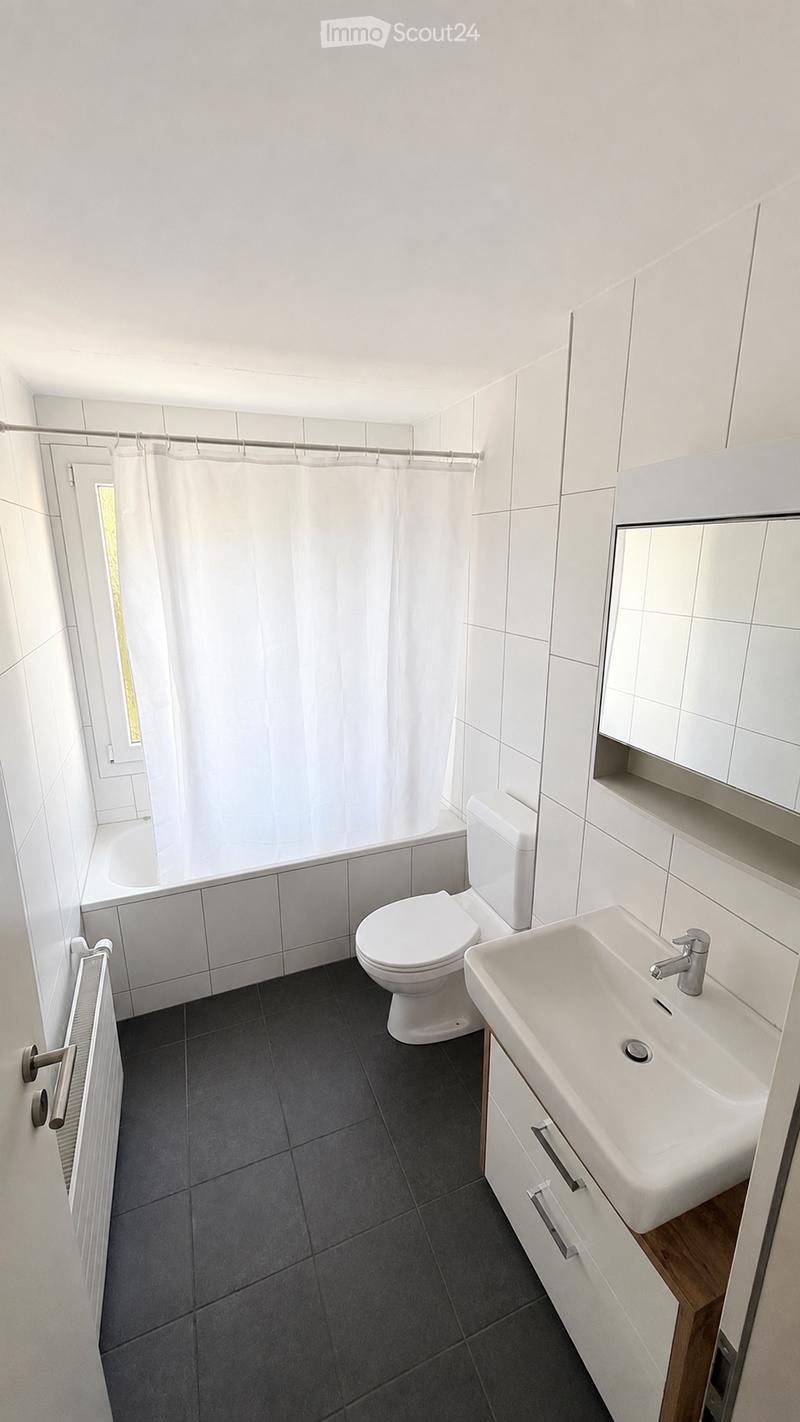 Modern bathroom with white tiles, dark grey floor, toilet, sink, bathtub, and shower curtain.