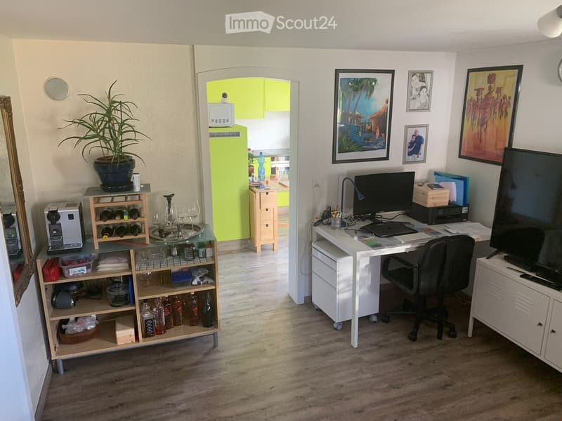 A well-furnished room with a desk, chair, laptop, screen, wine rack, and a small kitchen area visible in the background.