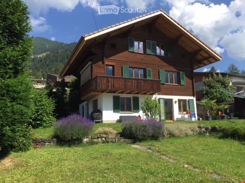 Two-story wooden house with green shutters, balcony, and well-maintained garden with lavender plants and a stone pathway.