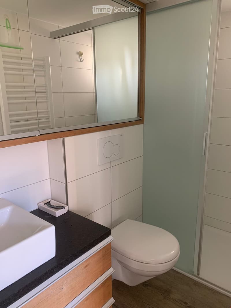 Modern bathroom with white tiles, a black countertop, a white sink, a toilet, a large mirror, and a glass-enclosed shower. A towel rack and a shaving kit are visible on the countertop.