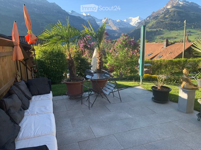 Outdoor patio with a white sofa, small table, chairs, potted plants, and a scenic mountain view.