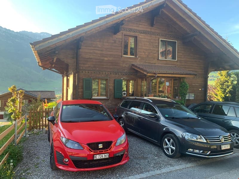 A large wooden house with multiple windows and a sloped roof, surrounded by a scenic mountain landscape. Three cars are parked in front of the house.