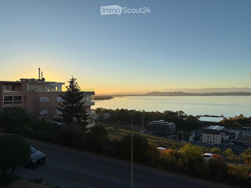 A modern multi-story building with balconies overlooking a serene lake and distant mountains during sunset.
