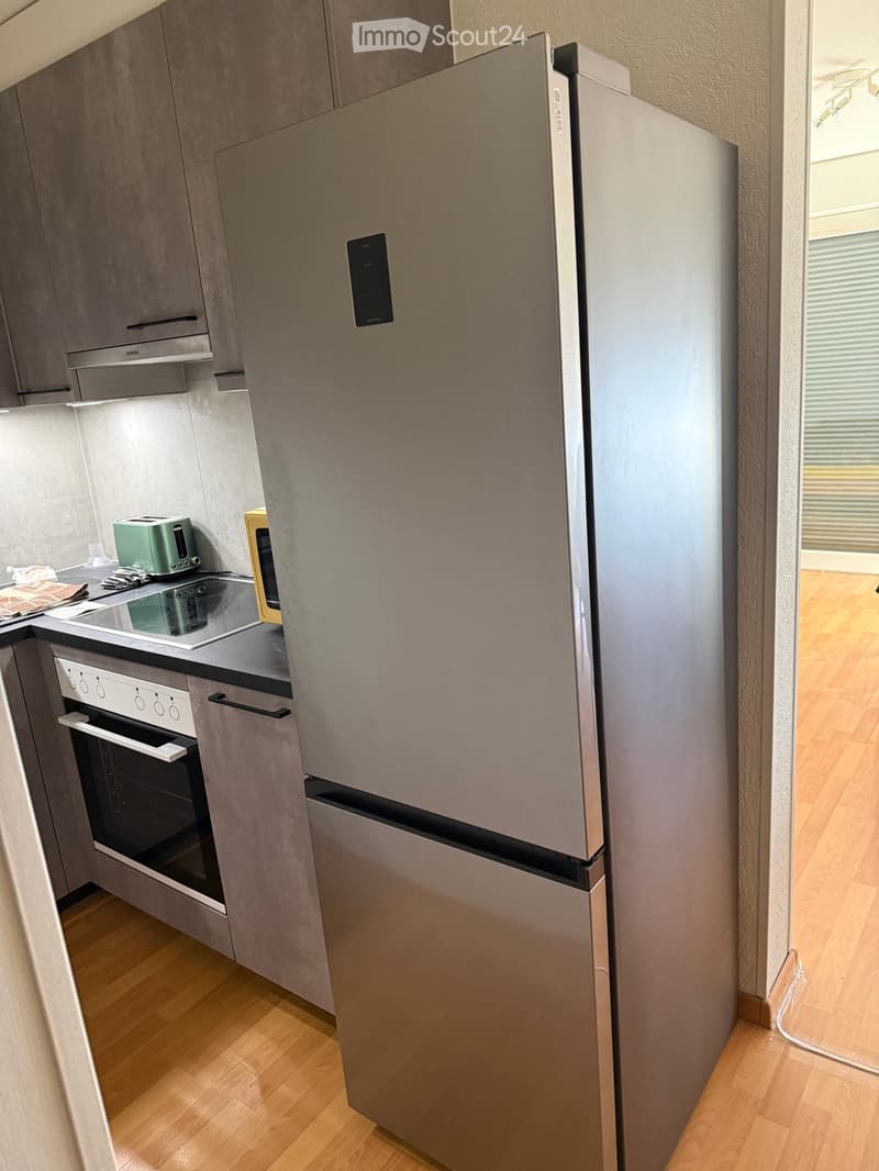 Modern kitchen with stainless steel appliances, including a refrigerator, oven, and toaster, set against grey cabinets and a wooden floor.