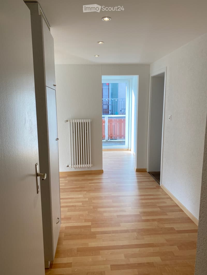 Empty hallway with wooden flooring, white walls, and recessed ceiling lights. A radiator is mounted on the left wall, and a door leads to a balcony area visible at the end.