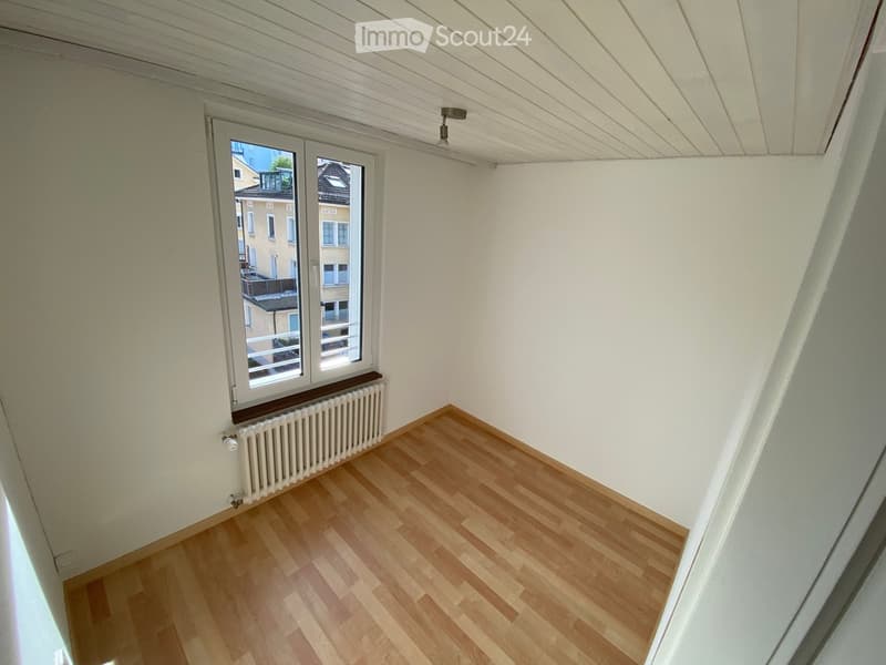 Empty room with wooden flooring, white walls, and a window with a radiator below it. The ceiling has a slanted design with a light fixture.