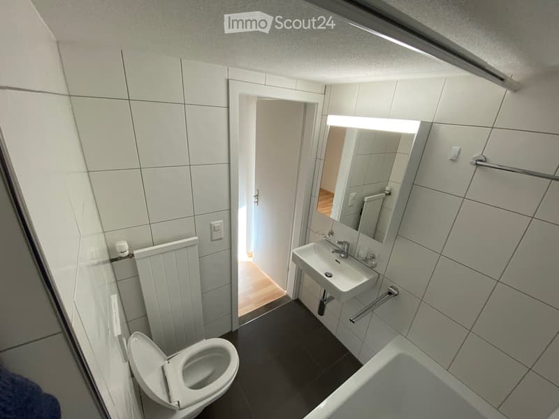 Modern bathroom with white tiled walls, a toilet, a sink with a mirror, a bathtub, and a radiator. The door to the adjacent room is open, revealing a wooden floor.