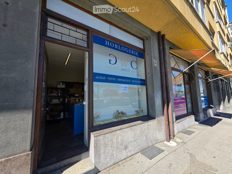 A storefront with a glass window displaying the sign "HORLOGERIE DWC GERZNER WATCH CENTRE" and the words "ACHAT - VENTE - REPARATION - RESTAURATION" along with a "À LOUER" sign. The interior shows some furniture and items, including a desk and chairs.