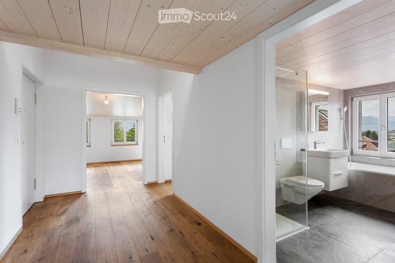 Modern bathroom with wooden flooring, white walls, and a glass-enclosed shower area. Features a toilet, sink, and bathtub, with large windows offering a scenic view.